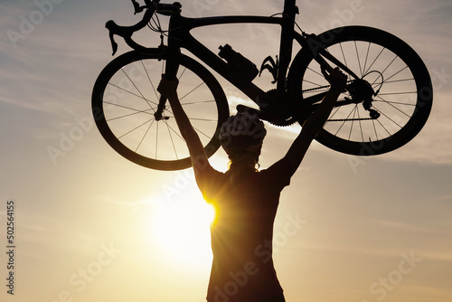 Female road cyclist raising bicycle with both arms above her head at sunset with beautiful green nature. Winning, success and achievement concept.