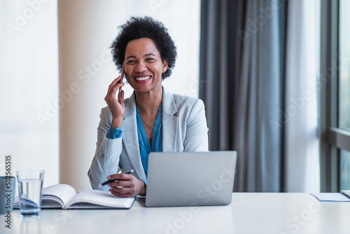 Portrait of happy businesswoman talking on cellphone while using laptop at desk