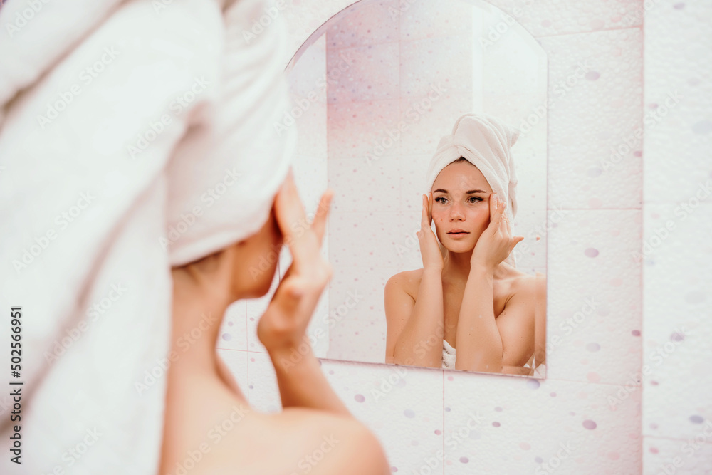 Young beautiful woman using face cream moisturizing lotion after bath. Pretty attractive girl in a towel on her head stands in front of a mirror in a home bathroom. Daily hygiene and skin care