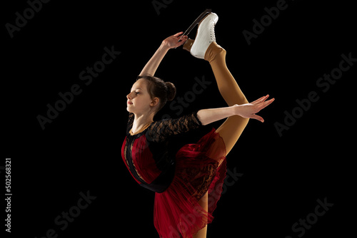 Studio shot of little female figure skater in beautiful stage attire skating isolated on black background in spotlight. Concept of movement, sport, beauty.