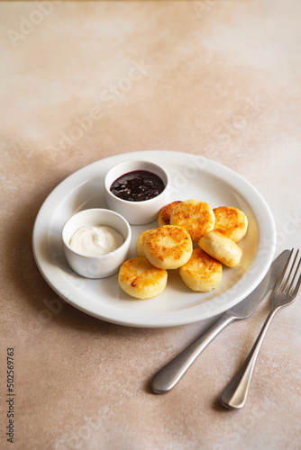 Cottage cheese pancakes, syrniki, ricotta fritters on white ceramic plate with sour cream and jam. Healthy and delicious morning breakfast. Beige background. Food for kids. Ukrainian food.