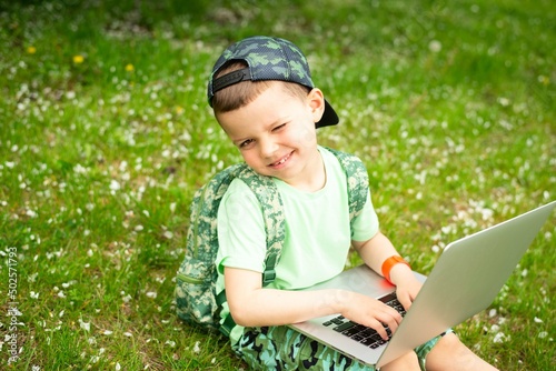 A cheerful kid with a laptop sits on green grass in green clothes and a cap.The boy is typing text on the computer. © volkonskaya