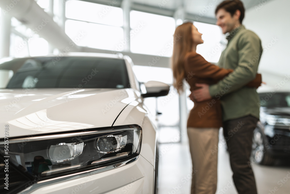 Happy young couple hugging at auto dealership, celebrating purchase of new vehicle, selective focus on car
