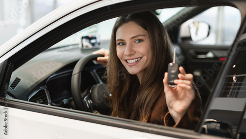 Cheerful young woman holding key from auto, buying new car at showroom, sitting inside automobile salon at dealership