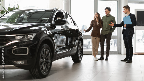 Confident car salesman showing new car to millennial couple at auto dealership, banner design © Prostock-studio