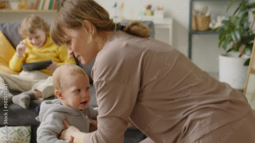 Happy mother supporting baby son as he making his first steps at home, lifting him up and smiling while elder kid playing on tablet on sofa