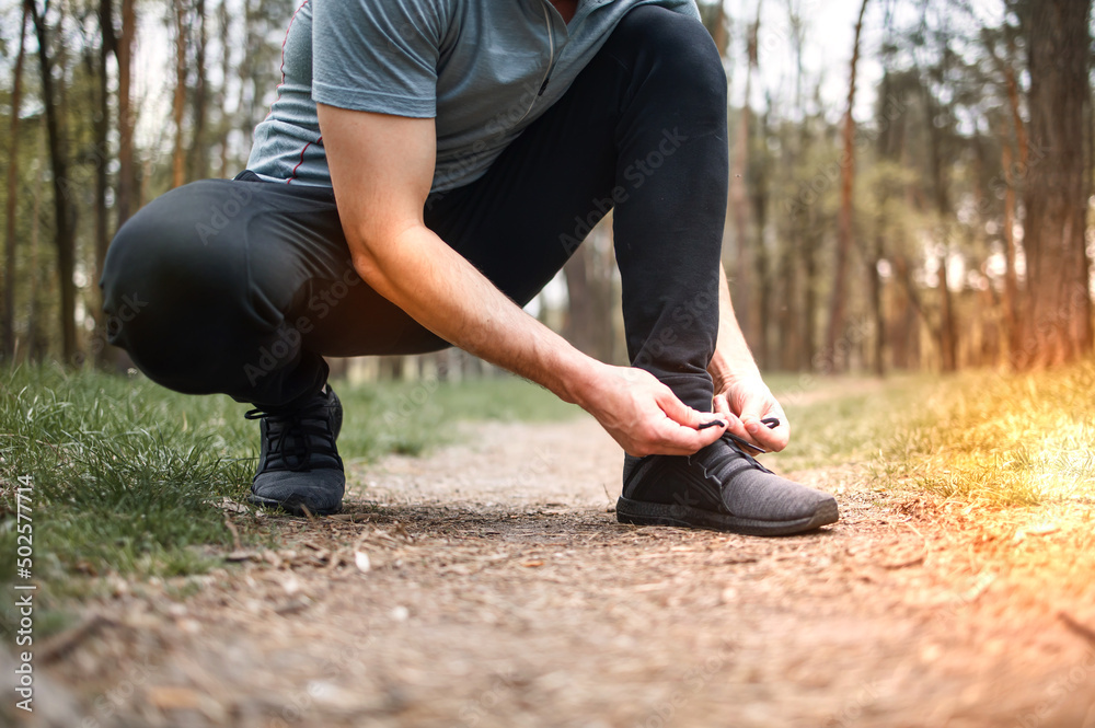 Man tying sneakers. Male runner jogging at the park. Guy training outdoors. Exercising on forest path. Healthy, fitness, wellness lifestyle.