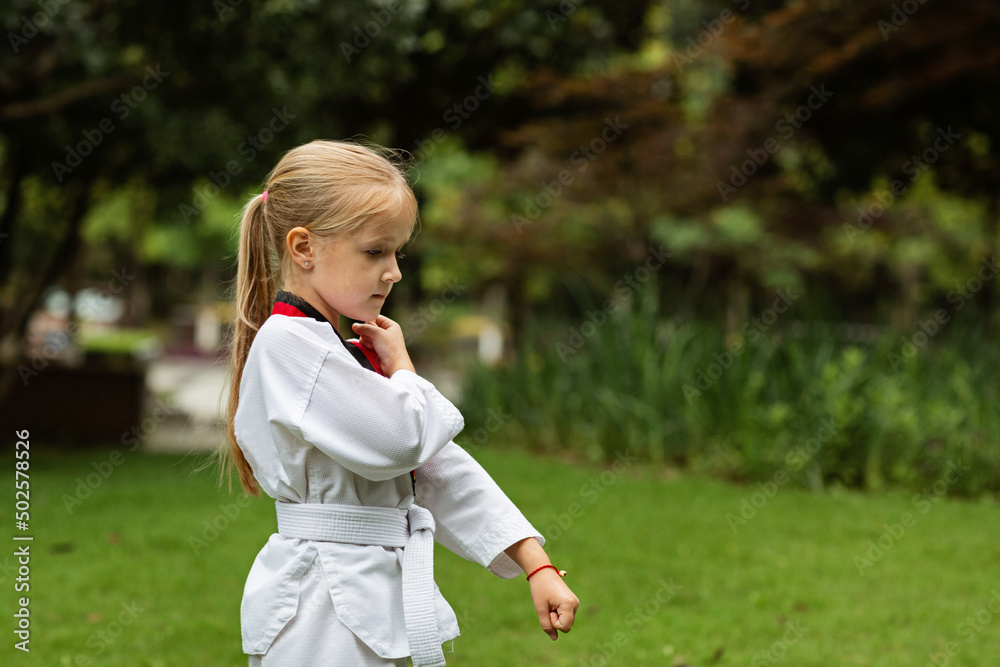 Caucasian little girl six years old in kimono with white belt exercising Taekwondo at summer park alone during coronavirus covid-19 lockdown, self isolation and social distancing Caucasian little girl six years old in kimono with white belt exercising Taekwondo at summer park alone during coronavirus covid-19 lockdown, self isolation and social distancing