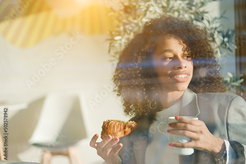 Young smiling african american girl  drinking coffee and eating tasty croissant in a cozy cafe. Holidays  lunch time and vacation concept 