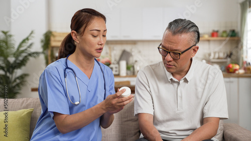 asian womale caregiver explaining medicine prescription to senior man patient during home visit. she holds a drug bottle and talking with hand gestures