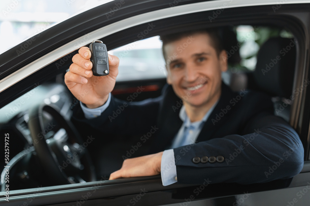 Successful millennial salesman showing car key from auto window at modern dealership, selective focus