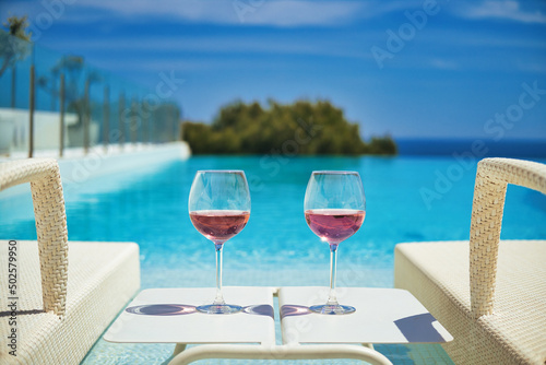 Foto Two glasses with cocktails on table between deck chairs sunbeds on the swimming pool, sea and blue sky background