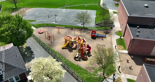 Elementary school recess for students. Children run and play on playground equipment. Beautiful spring aerial with blooming trees.