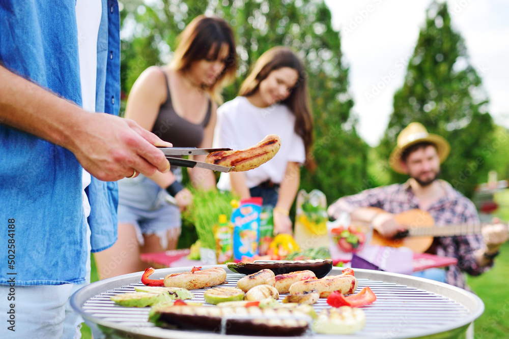 a man's hand holds a barbecue tongs with a juicy delicious meat steak against the background of a barbecue grill with meat and vegetables and a group of friends on a picnic who are having fun and