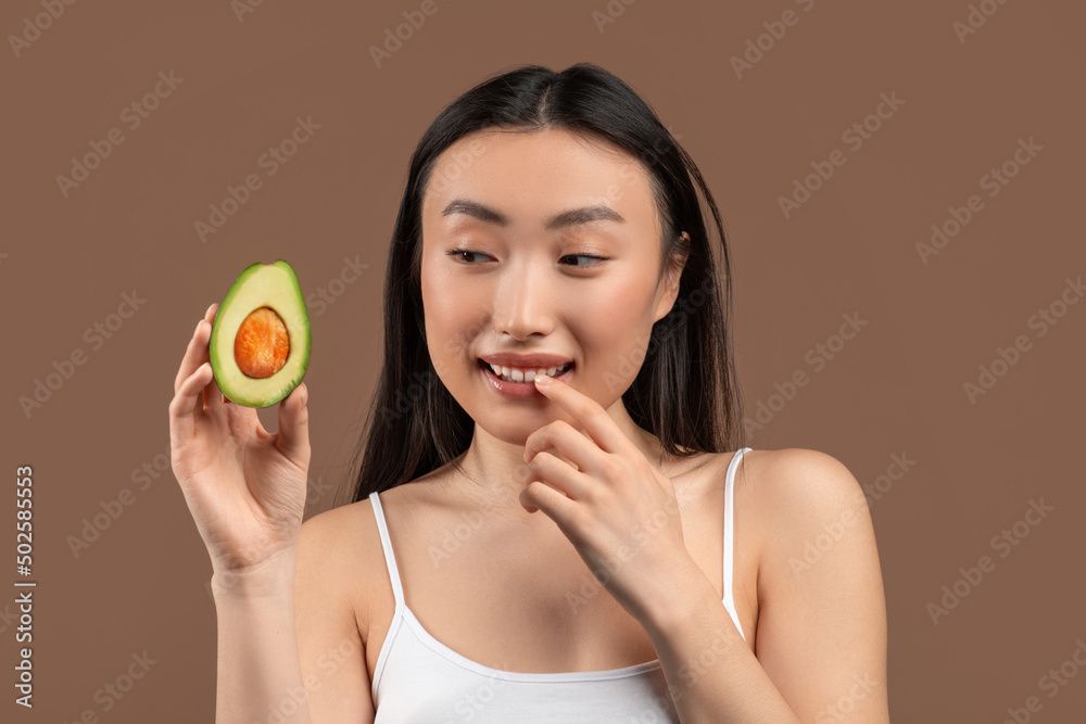 Natural skincare concept. Young asian woman holding ripe fresh avocado half, standing on brown background Natural skincare concept. Young asian woman holding ripe fresh avocado half, standing on brown background