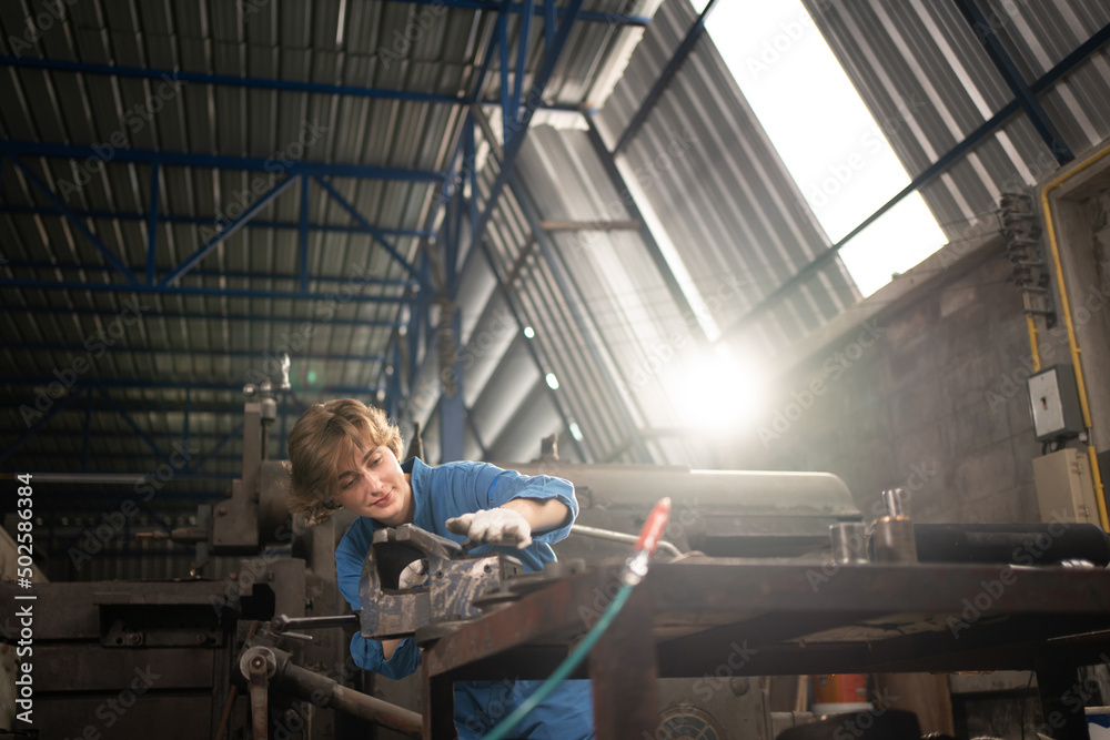 Low angle view selective focus of a Caucasian female lathe worker in a blue uniform and gloves, standing at a table with tools, working with a flat iron rod on a bench vise in a metal industry factory