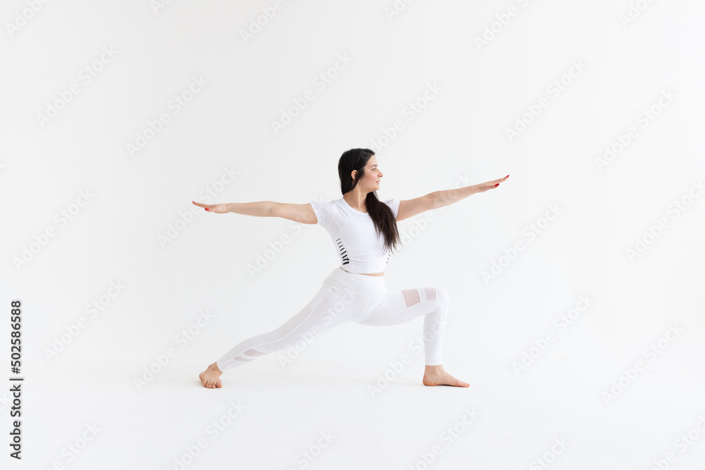 a beautiful young girl with dark hair stands in the pose of Virabhadrasana 2 on a white background. Yoga class