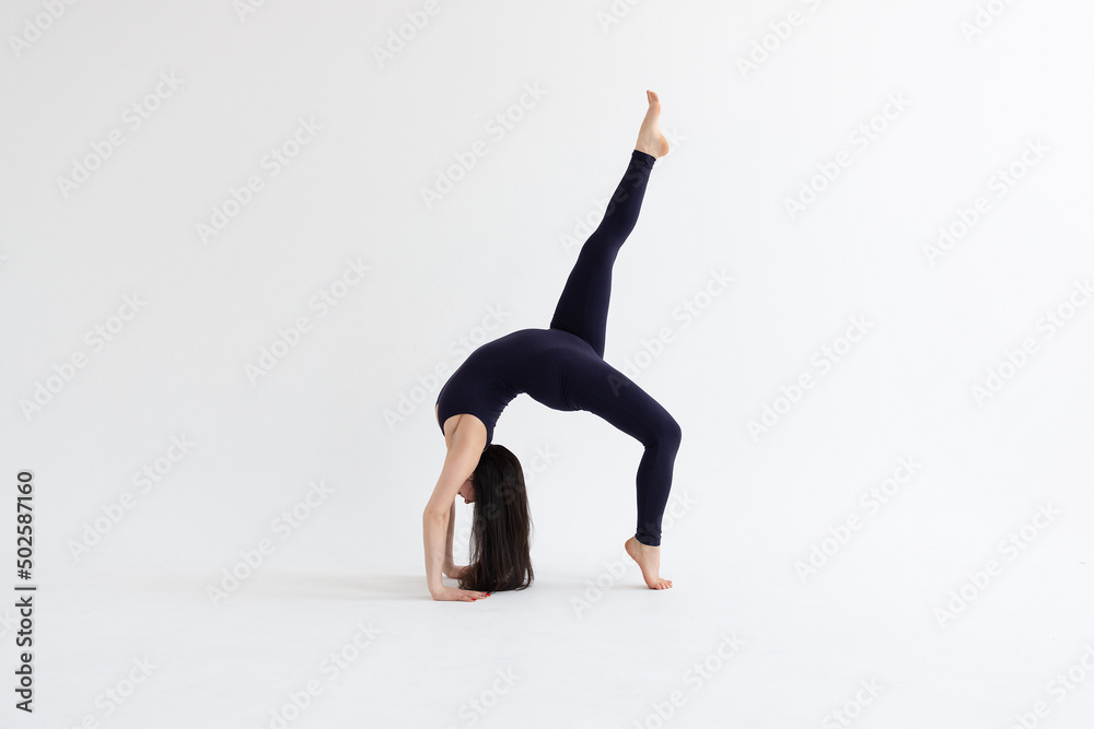 a beautiful young girl with dark hair stands in the pose of Eka Pada Urdhva Dhanurasana on a white background. Yoga class