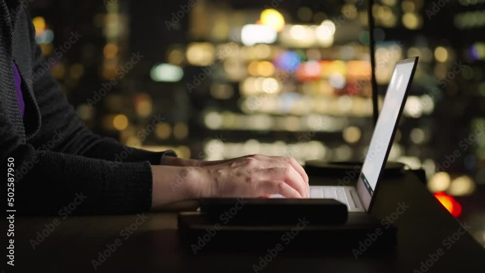 Motorized dolly shot of using laptop computer at night, with out focus city skyline background
