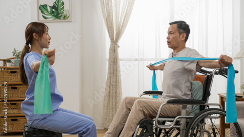 asian woman physiotherapist teaching handicapped older man how to use elastic band while she is doing resistance training on wheelchair at home