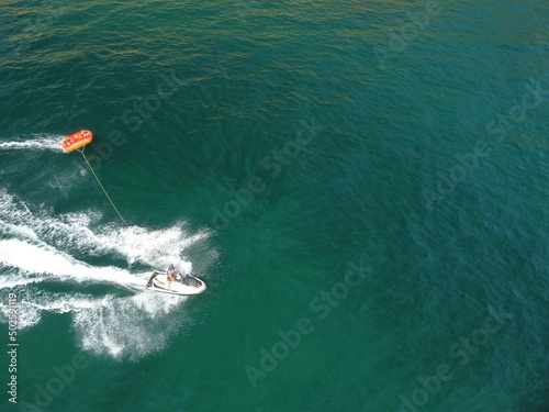 Happy people swim on air mattress behind a high speed water bike. Tourists ride on inflatable watercraft boat. Sea attraction. Water scooter skating people on large Inflatable raft. Aerial copter view