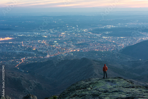 Wallpaper Mural Close up tourist, climber with dark red jacket standing on the top of Balkan mountain,staring at city Sliven at sunset. The shining lights of the city are sparkling in the far distance. Sport, travel Torontodigital.ca