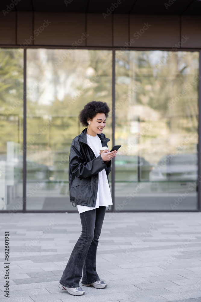 full length of smiling african american woman in earphone holding paper cup and using smartphone outside full length of smiling african american woman in earphone holding paper cup and using smartphone outside