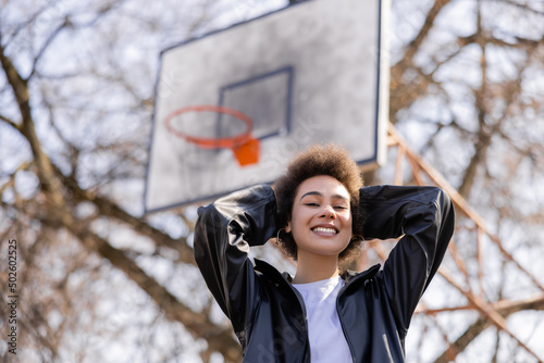low angle view of cheerful african american woman in jacket standing on basketball outdoor court