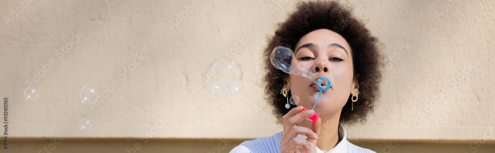 curly african american woman blowing soap bubble near wall, banner curly african american woman blowing soap bubble near wall, banner