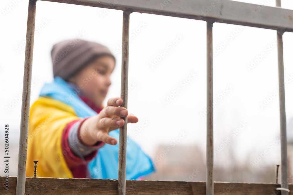 Obraz premium A little refugee girl with a sad look behind a metal fence. Social problem of refugees and internally displaced persons. Russia's war against the Ukrainian people