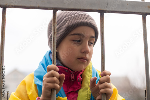 A little refugee girl with a sad look behind a metal fence. Social problem of refugees and internally displaced persons. Russia's war against the Ukrainian people photo