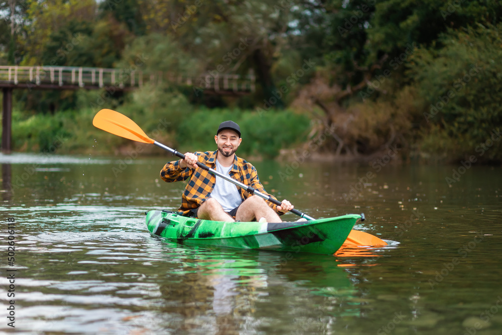 Fototapeta premium Happy bearded caucasian man in shirt and cap kayaking at the river with splashes. The concept of the World Tourism Day