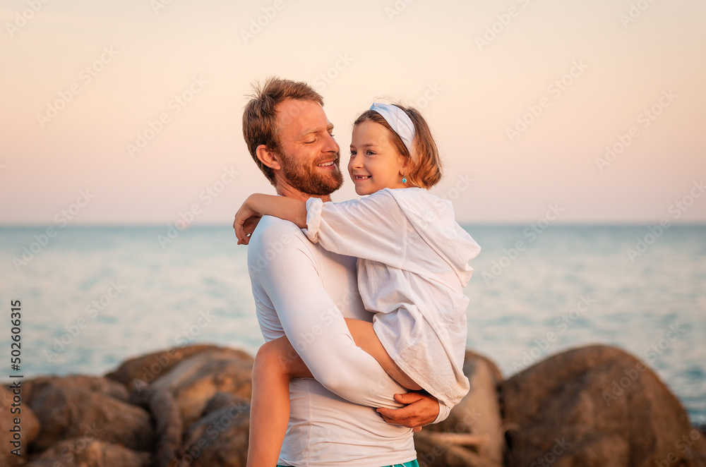 Father's Day. Smiling dad hugging daughter in his arms. In the background ocean and sky. Concept of happy fatherhood, parenting and adoption of children