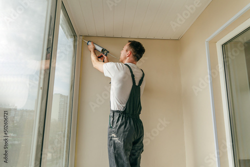 Φωτογραφία Young man wearing overalls sealing cracks between window and trim