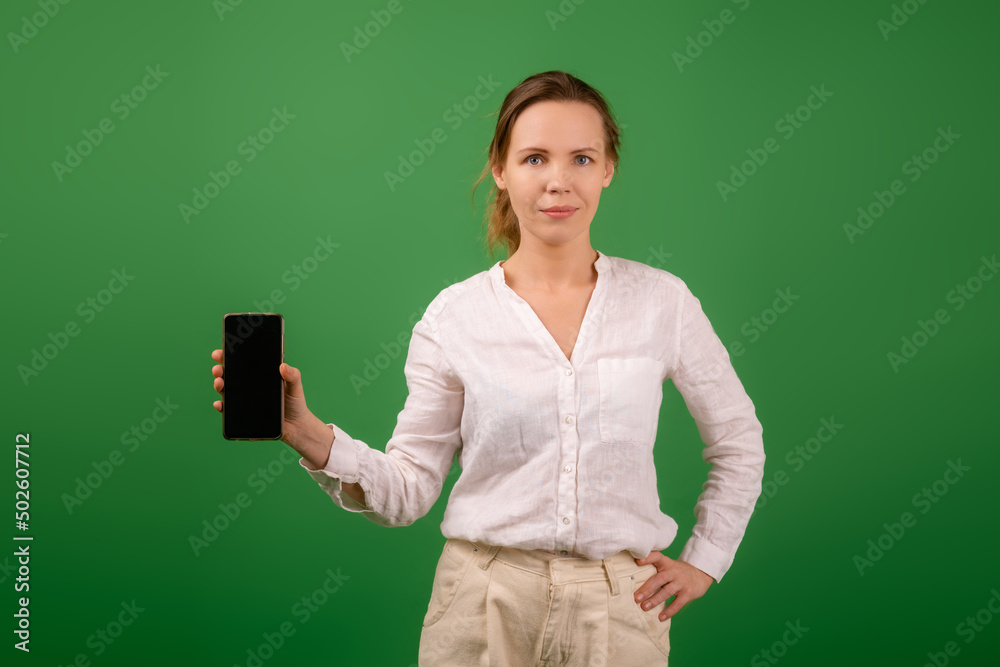 Attractive middle aged woman in a white shirt smiles and holds a mobile phone with the screen facing the camera on green. Mockup. Attractive middle aged woman in a white shirt smiles and holds a mobile phone with the screen facing the camera on green. Mockup.