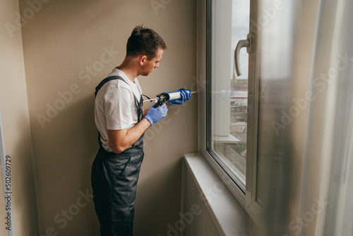 Young man wearing overalls sealing cracks between window and trim