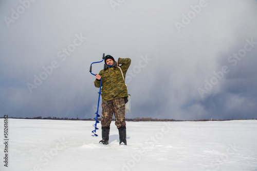A man - a fisherman stands on a lake with a drill and a box.