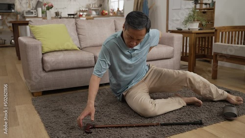 Asian elderly man trying to slowly get up after he slipped and fell on floor in the living room at home. he feels great pain in lower back while using stick to support himself