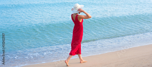 Young woman in red dress and white hat walking down the sand beach on sea summer vacation