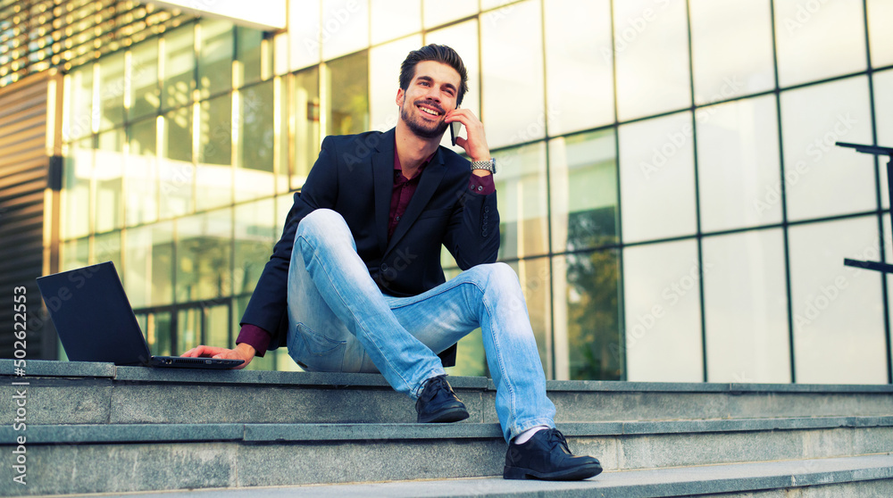 Young smiling businessman using phone and laptop while sitting on the stairs in front of corporate office building