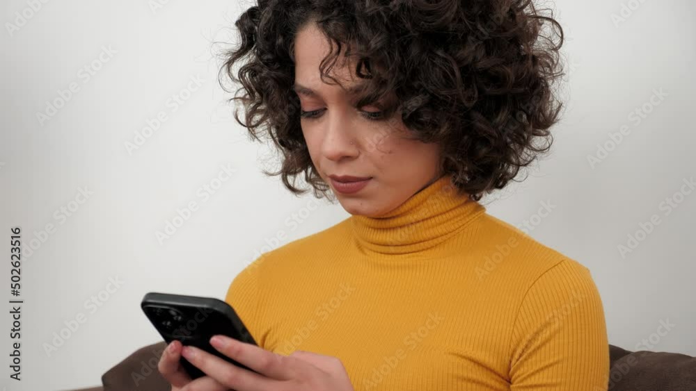 Hispanic curly woman student uses smartphone tapping screen display, sitting on couch at home. Businesswoman works for mobile phone online chatting with company employees, distance business meeting Hispanic curly woman student uses smartphone tapping screen display, sitting on couch at home. Businesswoman works for mobile phone online chatting with company employees, distance business meeting