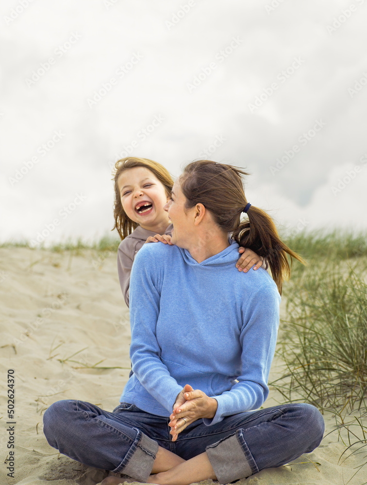 Youn mother sitting on the sand turning around and laughing with young daughter Youn mother sitting on the sand turning around and laughing with young daughter
