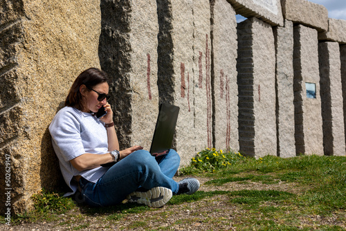 The girl sits on the grass near the rocks and works at the laptop. © Pablo Santos Somos