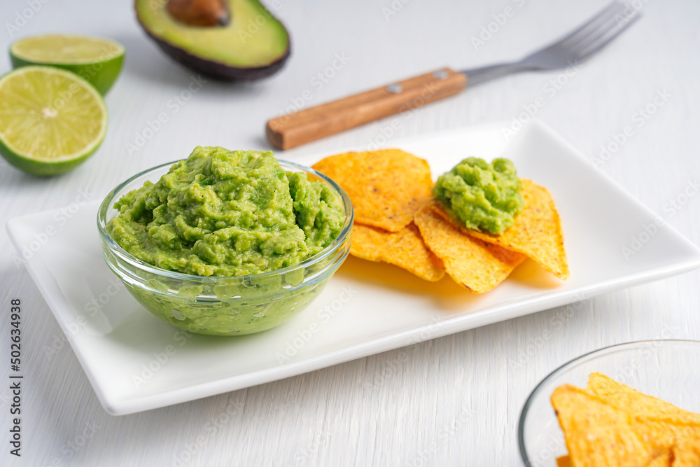 Mexican guacamole homemade vegan dip, spread, or salad made of mashed ripe green avocado served in glass bowl on plate with tortilla chips or nachos as appetizer on white wooden table with lime citrus Mexican guacamole homemade vegan dip, spread, or salad made of mashed ripe green avocado served in glass bowl on plate with tortilla chips or nachos as appetizer on white wooden table with lime citrus