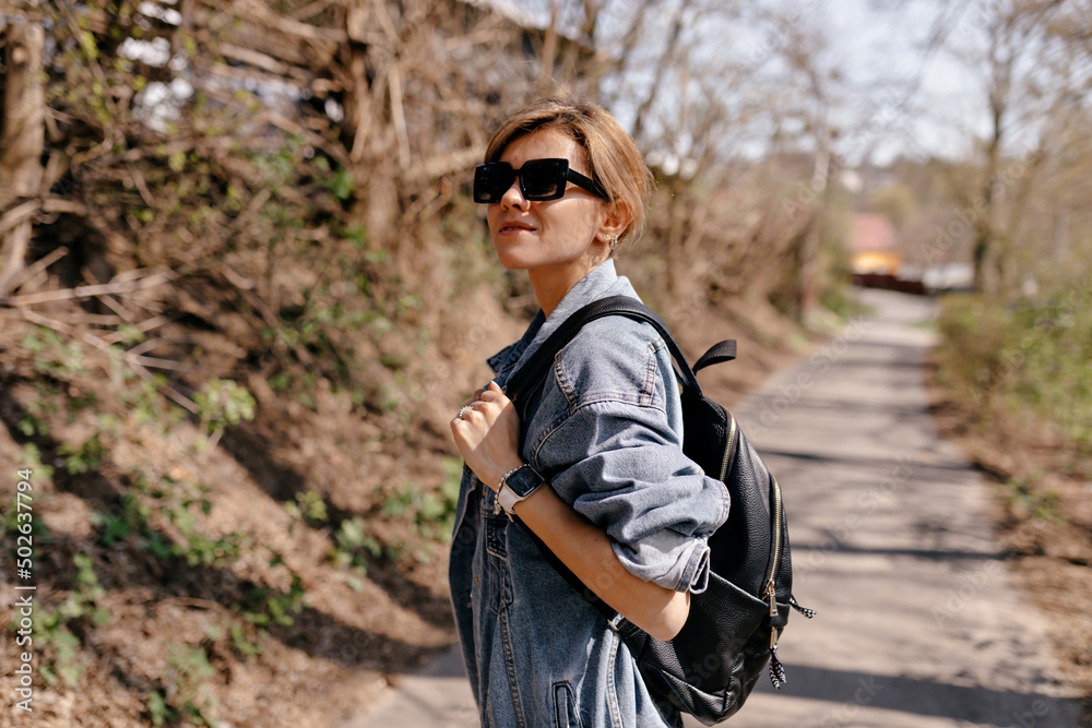 Pretty charming girl with light hair in sunglasses and denim jacket with backpack is walking on the forest trail in warm spring day