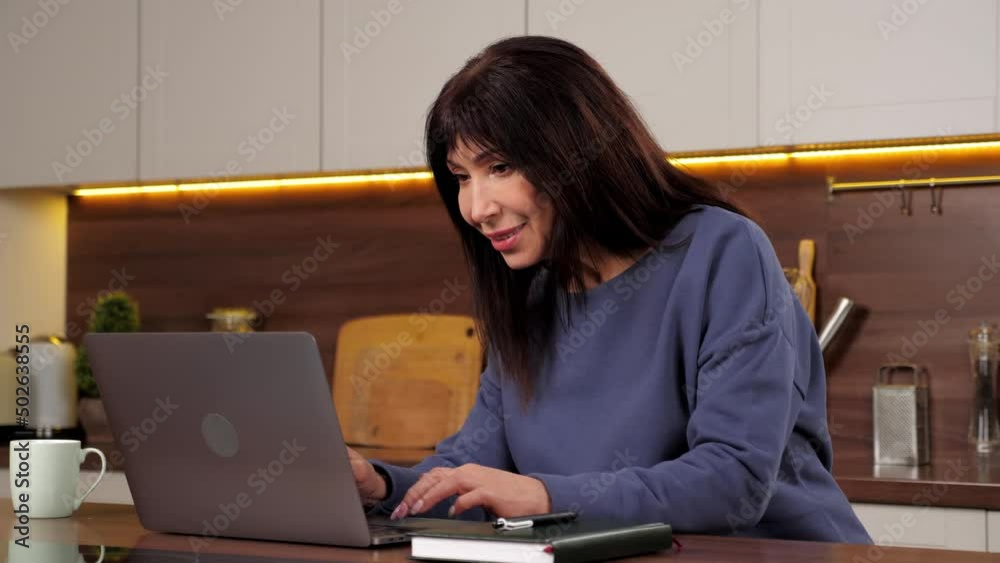 Smiling aged woman uses laptop typing text on keyboard in home kitchen. Positive businesswoman works for computer online chatting with company employees, distance meeting, sitting on chair at table Smiling aged woman uses laptop typing text on keyboard in home kitchen. Positive businesswoman works for computer online chatting with company employees, distance meeting, sitting on chair at table