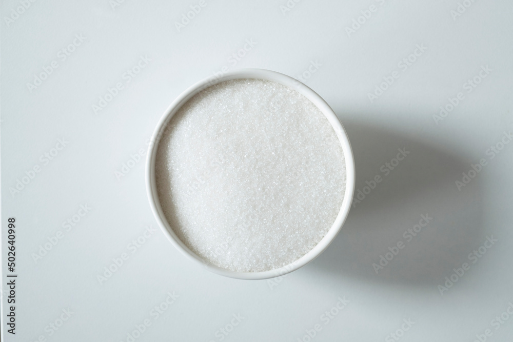 Granules of white sugar in a bowl on a white background Granules of white sugar in a bowl on a white background