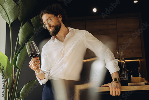 Curly man at home in a good kitchen holding a glass of red wine and looking at him. On the background of a beautiful strelitzia flower photo