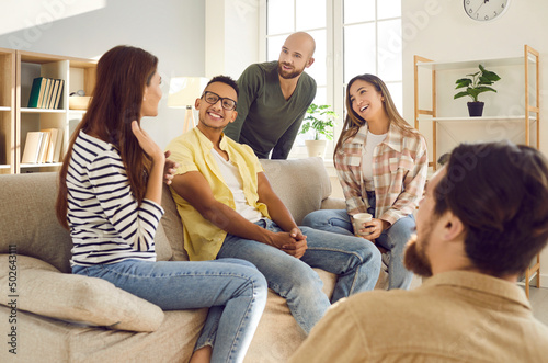 Spending great time with best friends. Group of young multiracial people have fun communicating with each other during meeting at home. Men and women tell their stories sitting on sofa in living room.