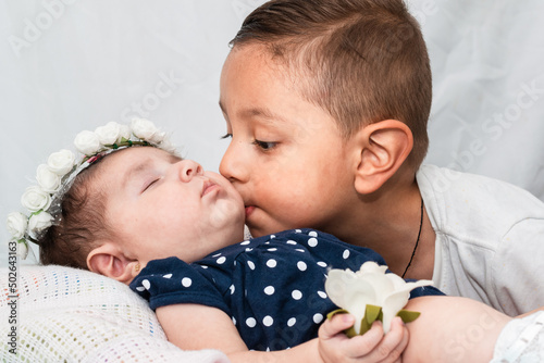 little latino boy kissing his little sister's cheek, the baby is lying on a white pillow, with her eyes closed, a crown of white roses and holding a rose. brother saying goodbye to his little sister. photo